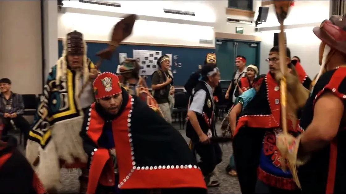 Several people in detailed red, black, and other regalia participating in an indoor group dance