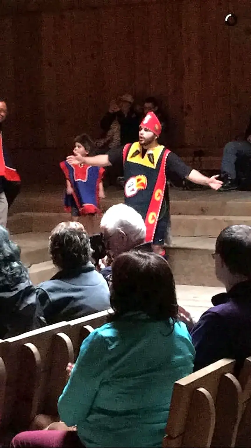 A person in red and black regalia speaking with arms extended at a gathering before an audience