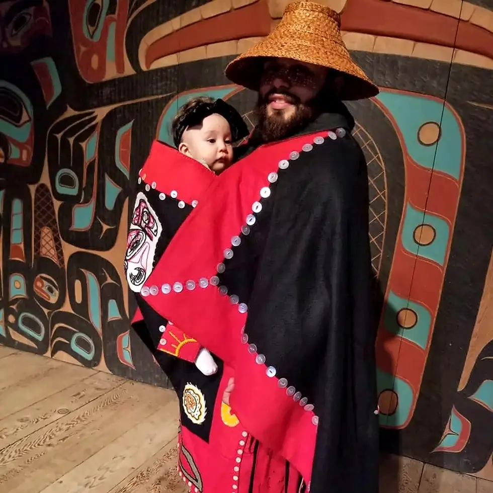 IM founder holding his child while wearing red and black regalia and a woven hat