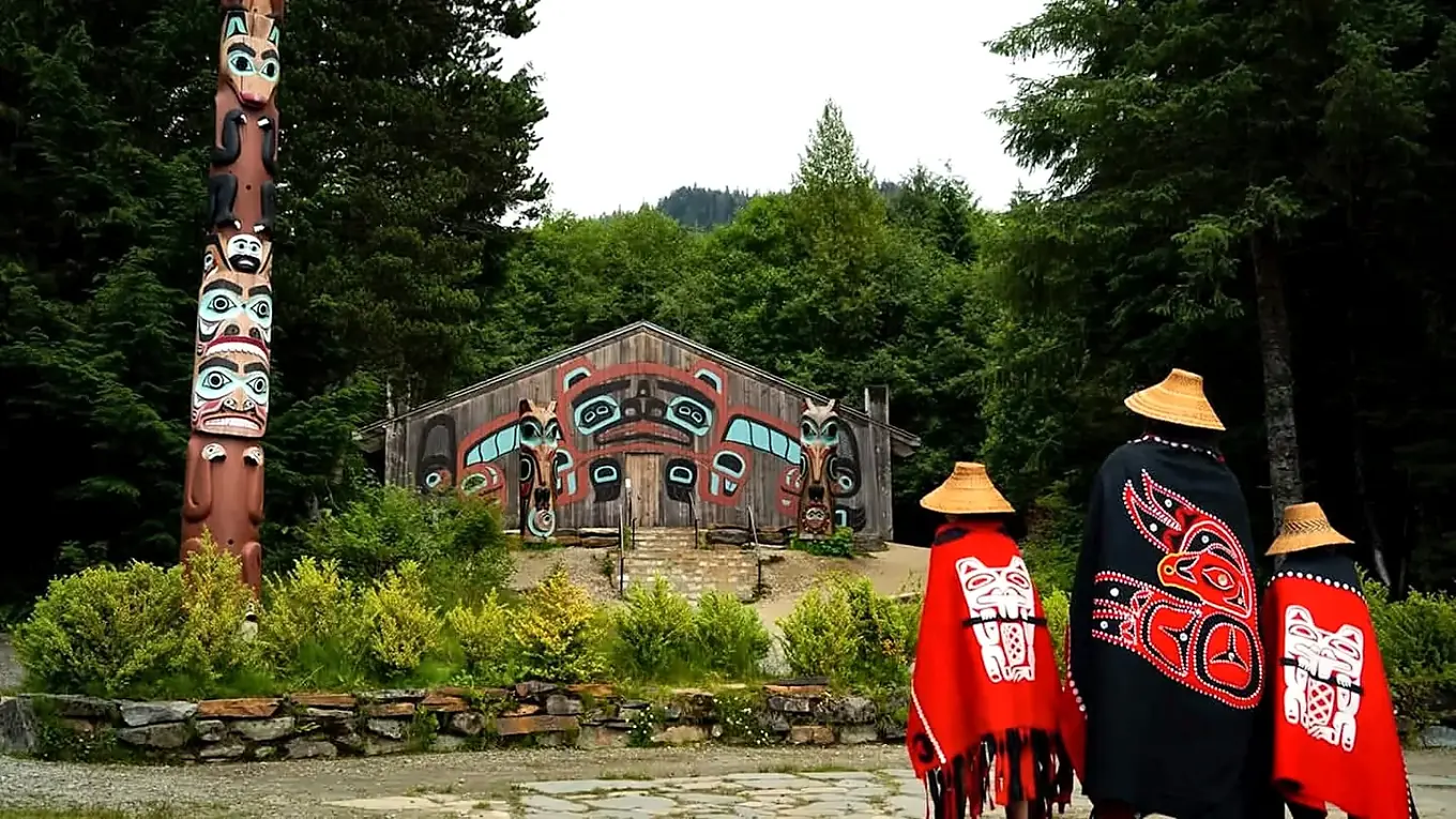 Three people wearing red and black regalia facing a traditional Northwest Coast-style longhouse and carved poles amid green forest