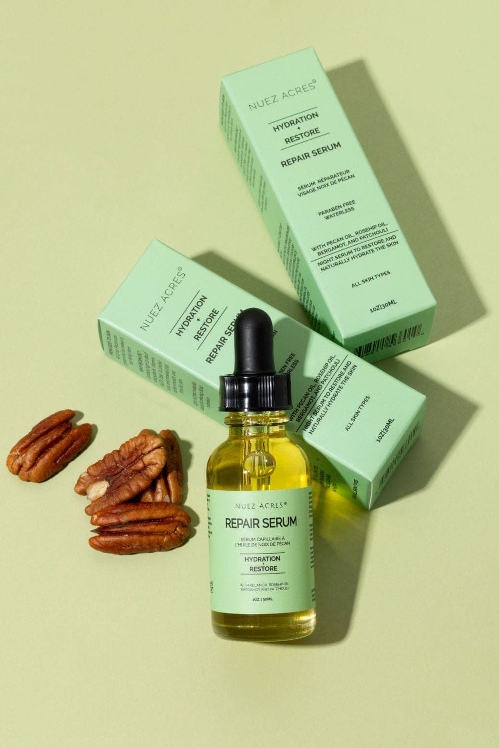 Nuez Acres Repair Serum bottle and green packaging on a pale green background beside pecans